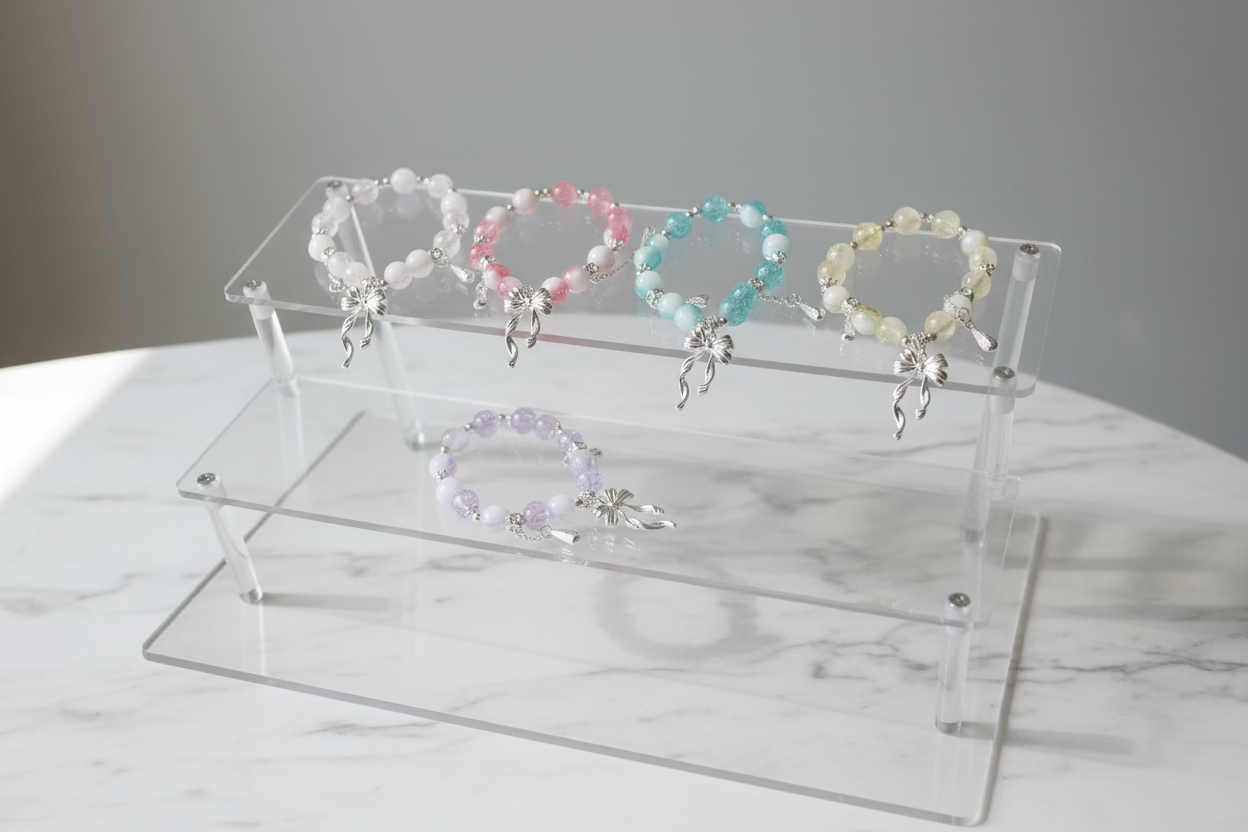 Five beaded bracelets with silver charms are displayed on a clear acrylic stand over a white surface. The bracelets feature pastel pink, light yellow, light blue, lavender, and white beads with silver bow charms.