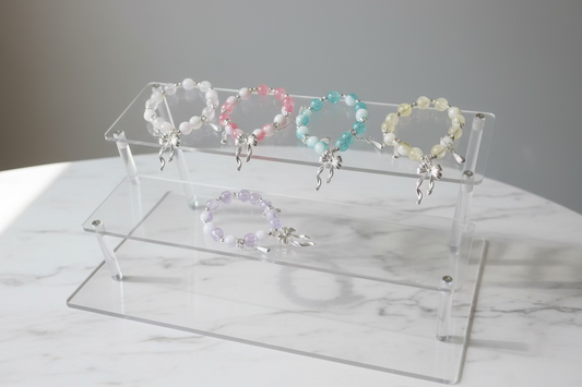 Five beaded bracelets with silver charms are displayed on a clear acrylic stand over a white surface. The bracelets feature pastel pink, light yellow, light blue, lavender, and white beads with silver bow charms.